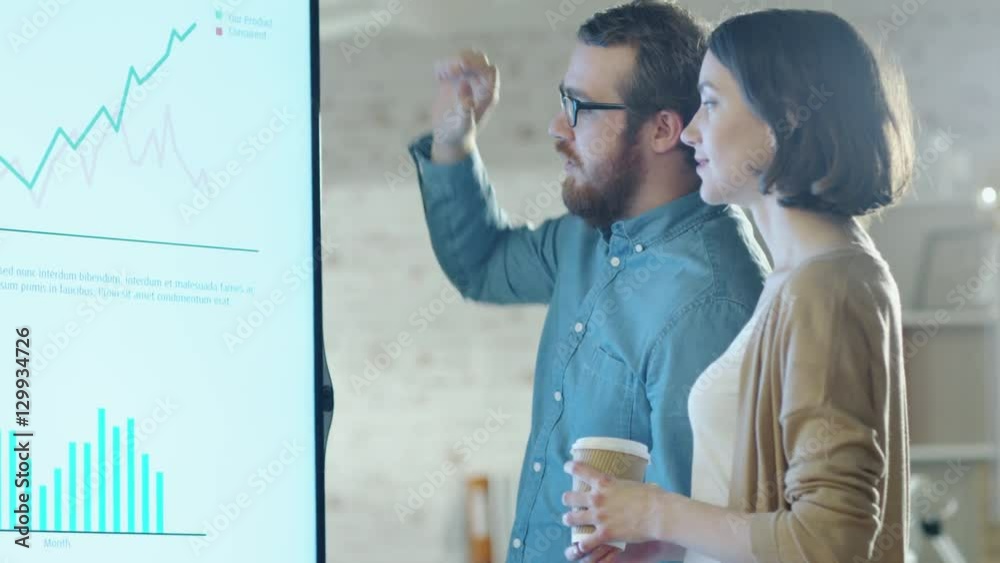 Young Man and Woman Discuss Charts Drawn on Their Electronic Whiteboard. Man Shows Details on the Screen Woman Listens Holding Cup of Coffee in Her Hands. Shot on RED Cinema Camera in 4K (UHD).