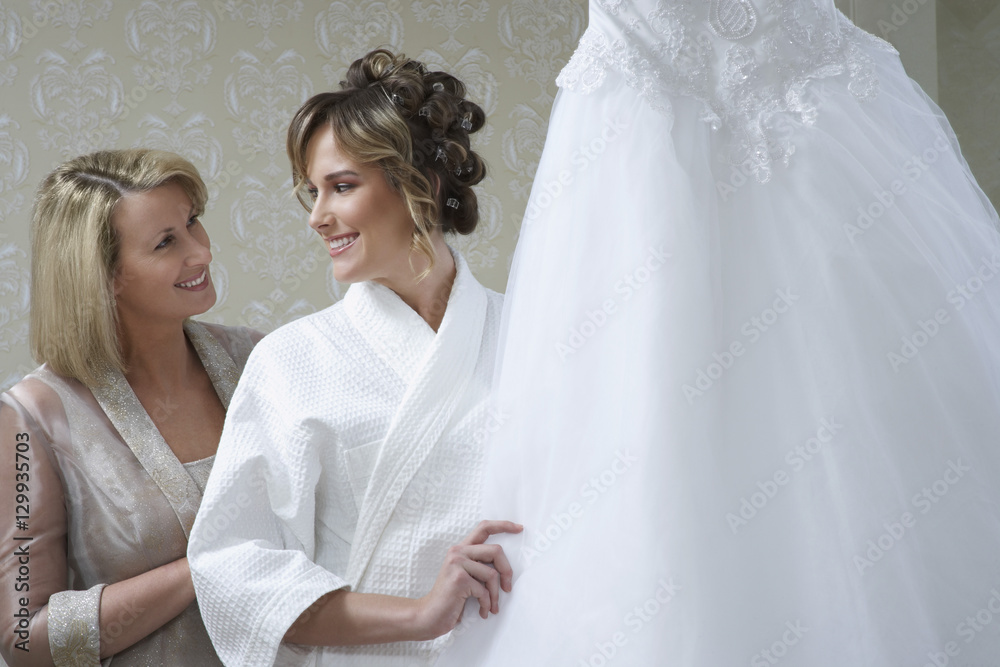 Happy mother with bride in bathrobe touching wedding dress