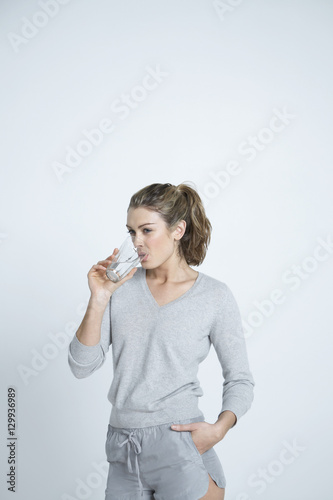 Beautiful young woman with hand in pocket drinking water from glass isolated over colored background