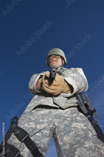 Low angle portrait of US army soldier aiming with pistol against blue sky