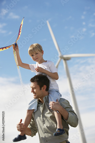 Little boy with kite sitting on father's shoulders at wind farm