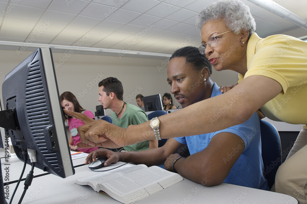 Senior teacher assisting male student during computer class with classmates in the background Senior teacher assisting male student during computer class with classmates in the background