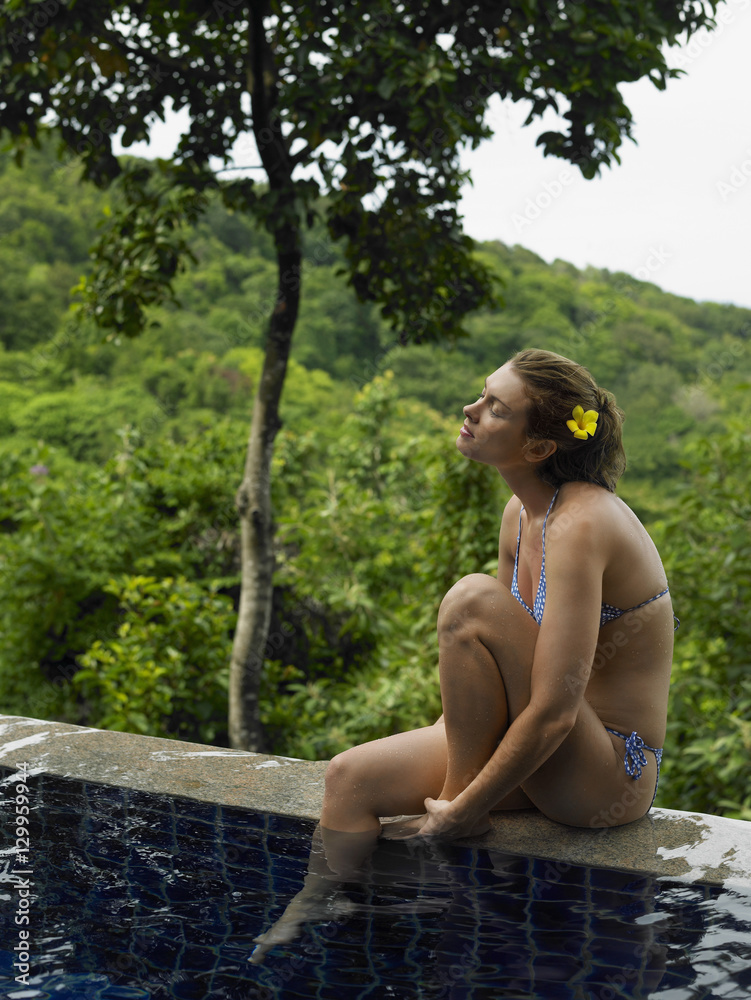 Fototapeta premium Side view of a bikini woman by swimming pool and against trees