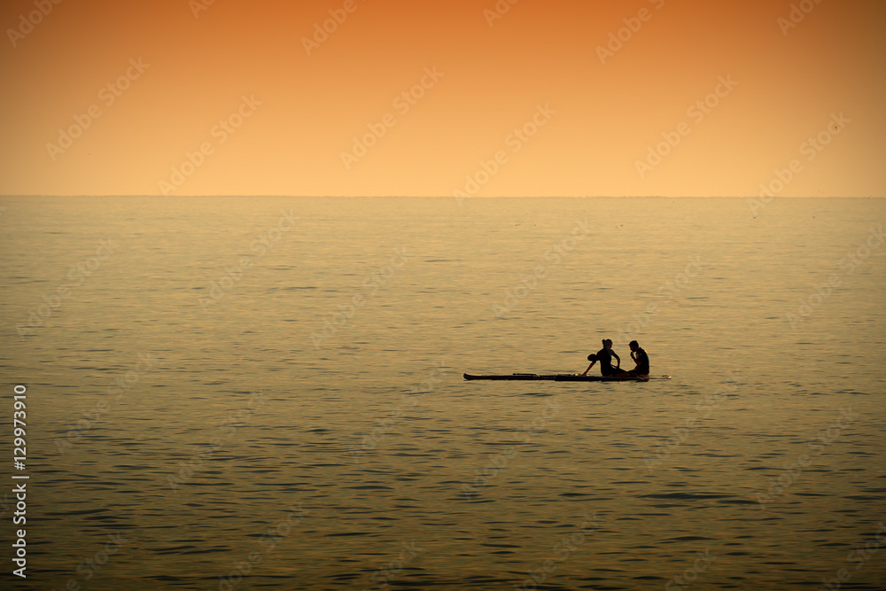 A couple resting while paddleboarding in a calm sea at sunset time. Dark orange atmosphere. Empty copy space for Editor's text.