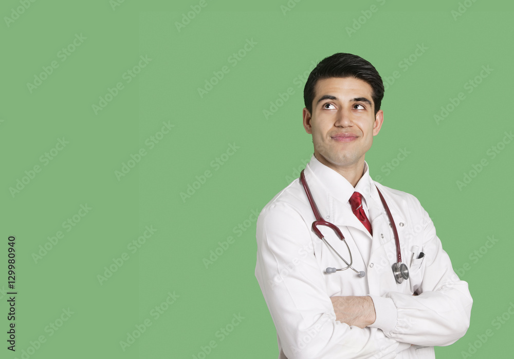 Male doctor in lab coat standing with arms crossed looking up over green background Male doctor in lab coat standing with arms crossed looking up over green background