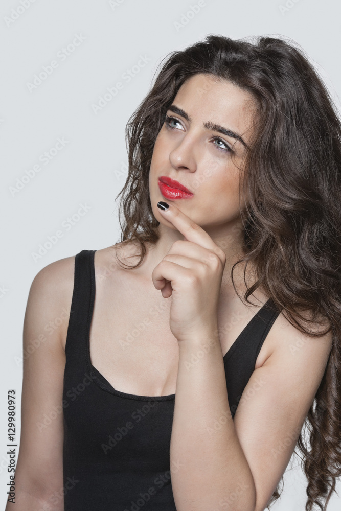 Thoughtful young woman looking away with hand on chin over gray background Thoughtful young woman looking away with hand on chin over gray background