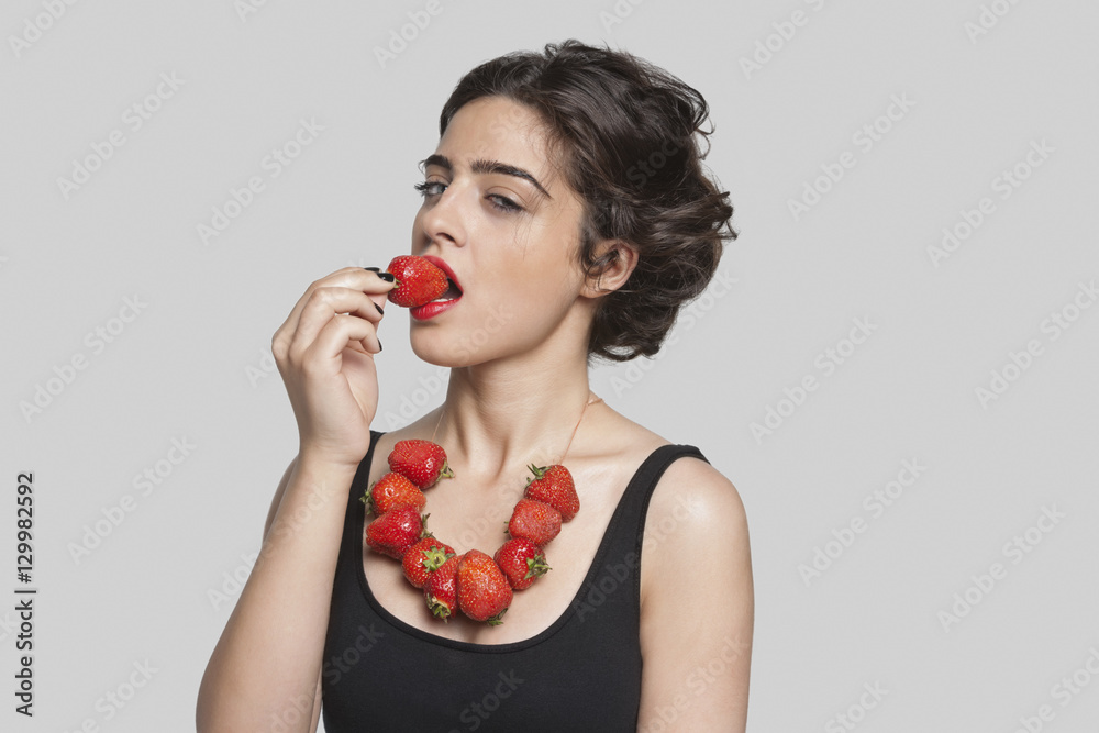 Seductive young woman wearing strawberry necklace as she eats one piece over gray background Seductive young woman wearing strawberry necklace as she eats one piece over gray background