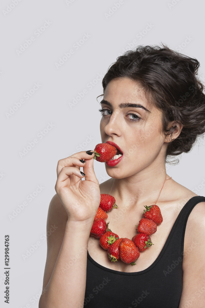 Portrait of a young woman wearing strawberry necklace as she eats one piece over gray background