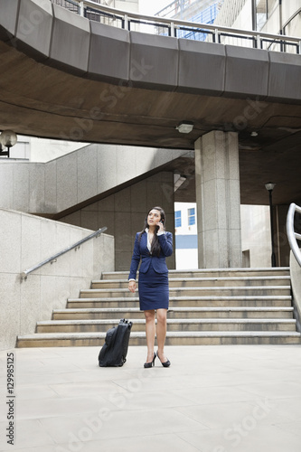Young Indian businesswoman using cell phone with luggage outdoors