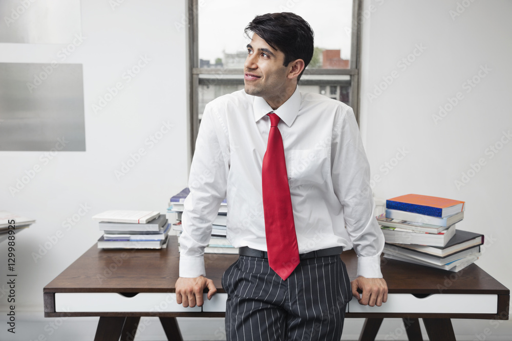 Indian businessman leaning on desk while looking away in office Indian businessman leaning on desk while looking away in office