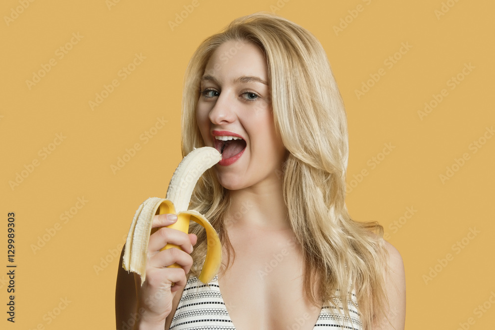 Portrait of a beautiful blond woman eating banana over colored background Portrait of a beautiful blond woman eating banana over colored background