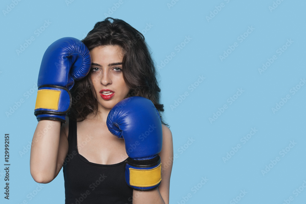 Portrait of an aggressive young woman wearing boxing gloves over blue background Portrait of an aggressive young woman wearing boxing gloves over blue background