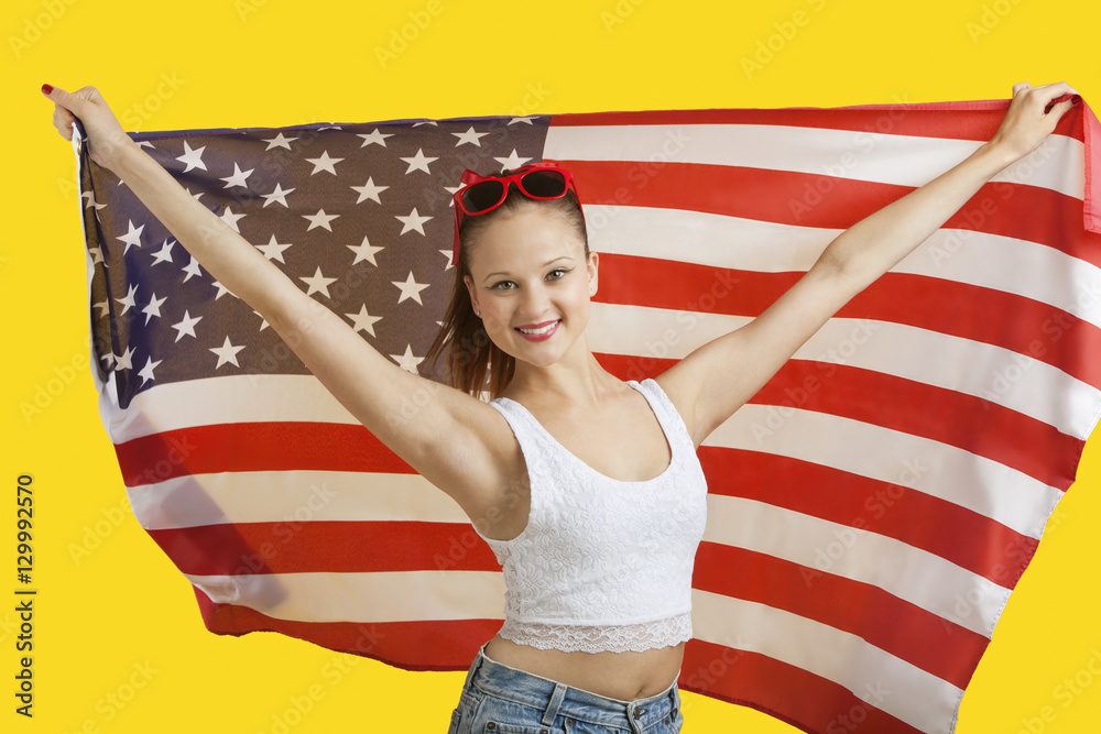 Fototapeta premium Portrait of happy young woman holding American flag over yellow background
