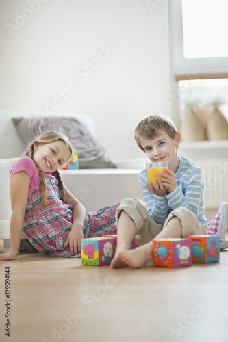 Portrait of young boy drinking orange juice while sitting with sister on floor