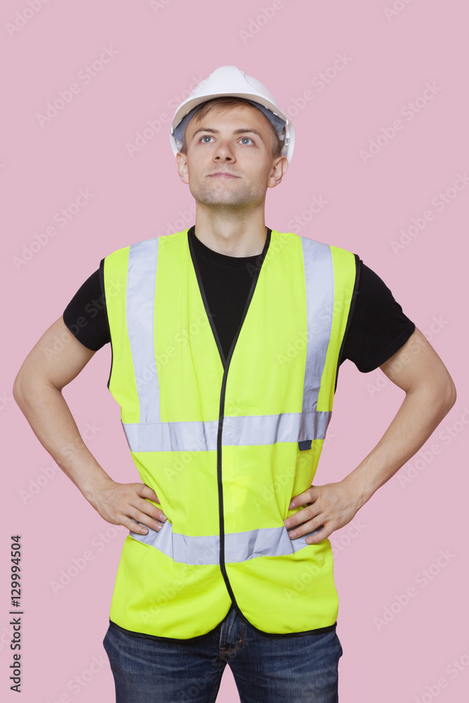 Handsome construction worker with hands on hips over pink background