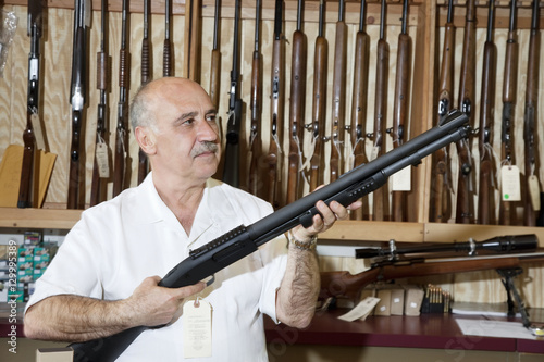 Mature gun store owner looking at weapon in shop