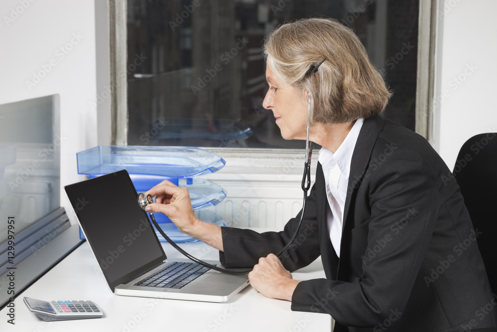 Side view of senior businesswoman examining laptop with the use of stethoscope at office desk Side view of senior businesswoman examining laptop with the use of stethoscope at office desk