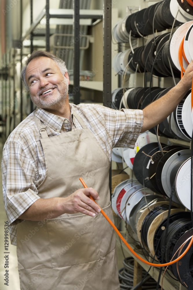 Fototapeta premium Happy mature salesperson standing by electrical wire spool while looking away in hardware store