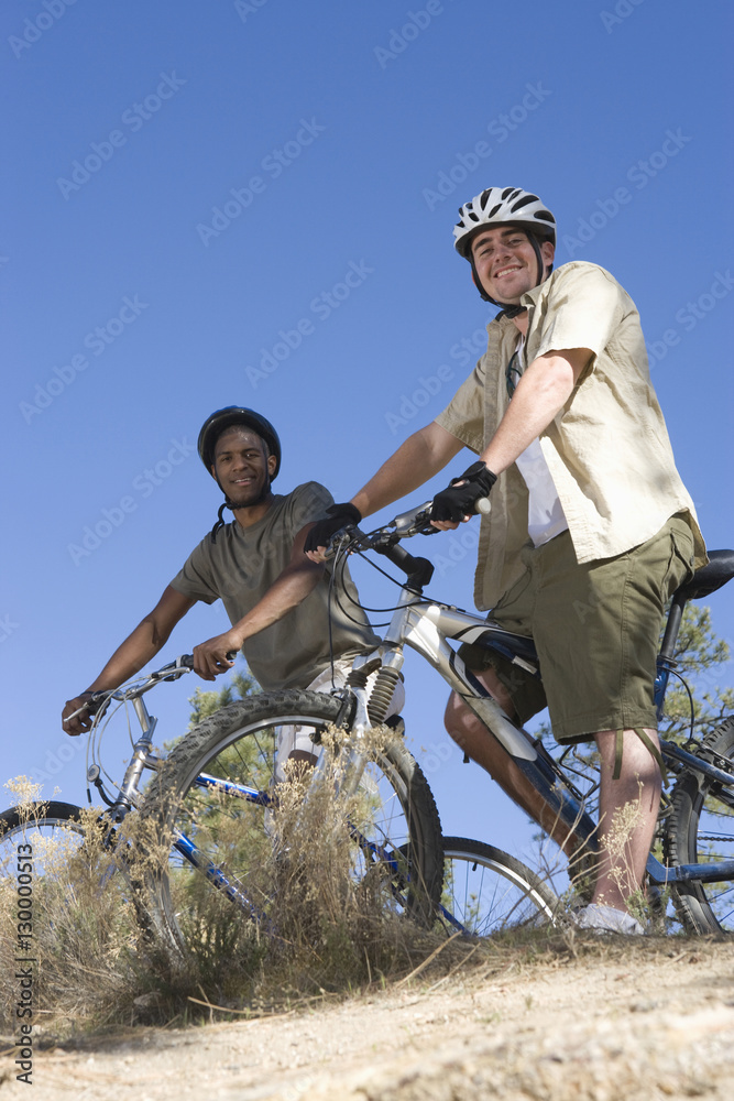 Low angle view of two multiethnic friends with mountain bikes on hilltop against clear blue sky Low angle view of two multiethnic friends with mountain bikes on hilltop against clear blue sky