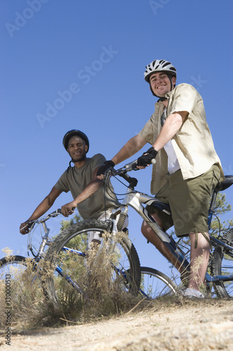 Low angle view of two multiethnic friends with mountain bikes on hilltop against clear blue sky