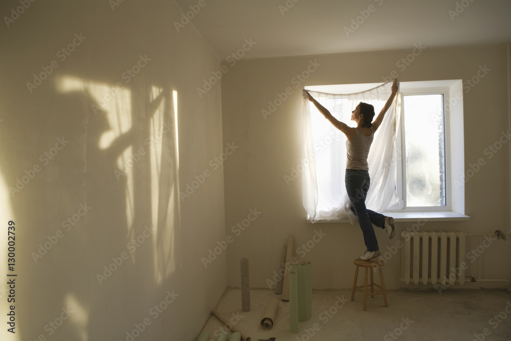Fototapeta premium Full length rear view of young woman fitting curtain in new apartment