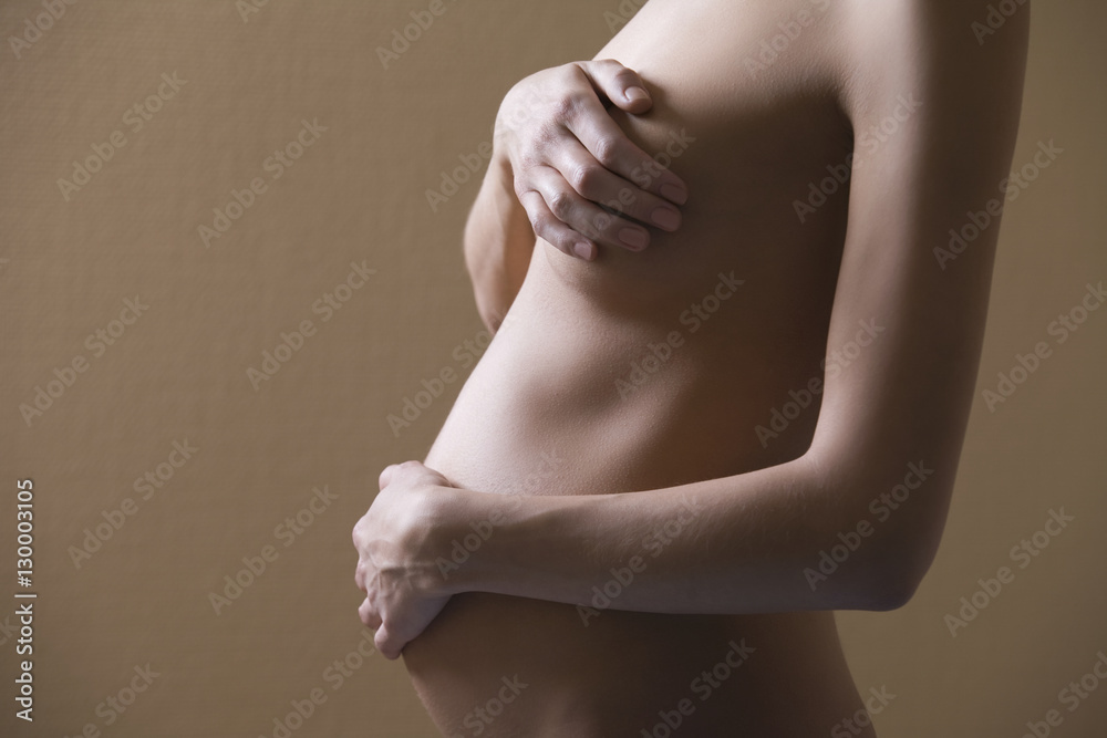Midsection side view of naked pregnant woman covering breast and abdomen on colored background