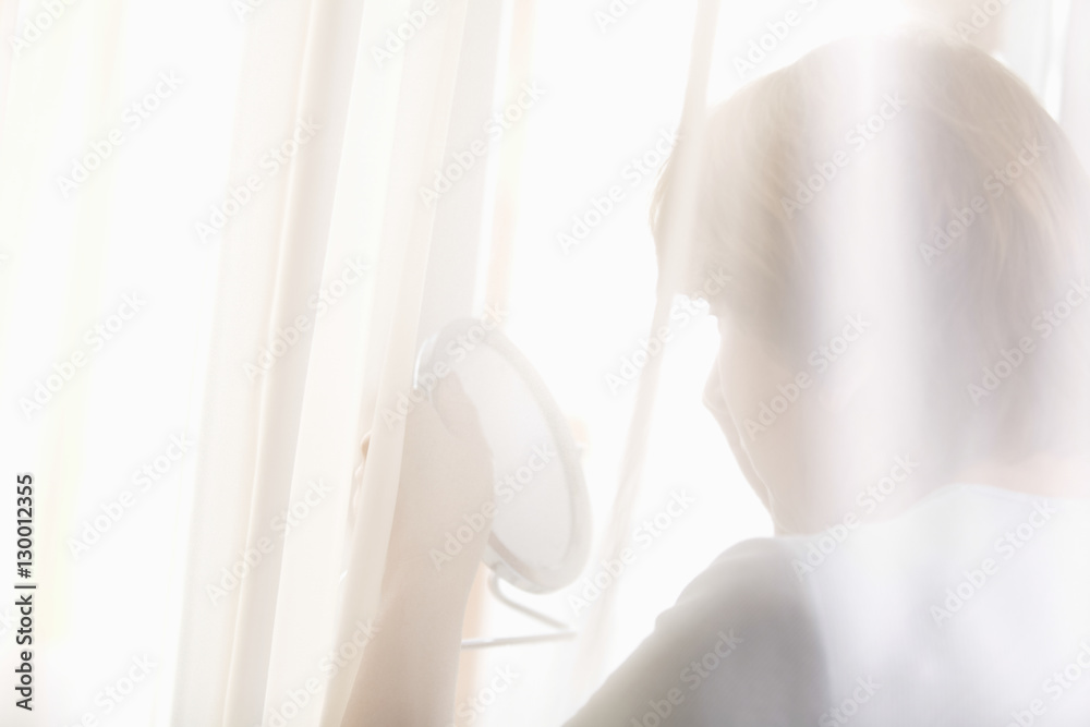 Rear view of senior woman examining herself in hand mirror behind curtains Rear view of senior woman examining herself in hand mirror behind curtains