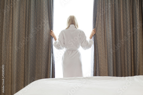 Rear view of young woman in bathrobe opening window curtains at hotel room