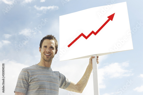 Young man looking away while holding arrow signboard against cloudy sky