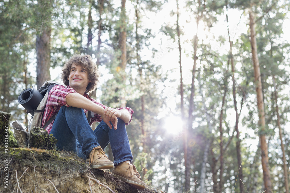 Full length of smiling male hiker looking away while sitting on cliff in forest Full length of smiling male hiker looking away while sitting on cliff in forest