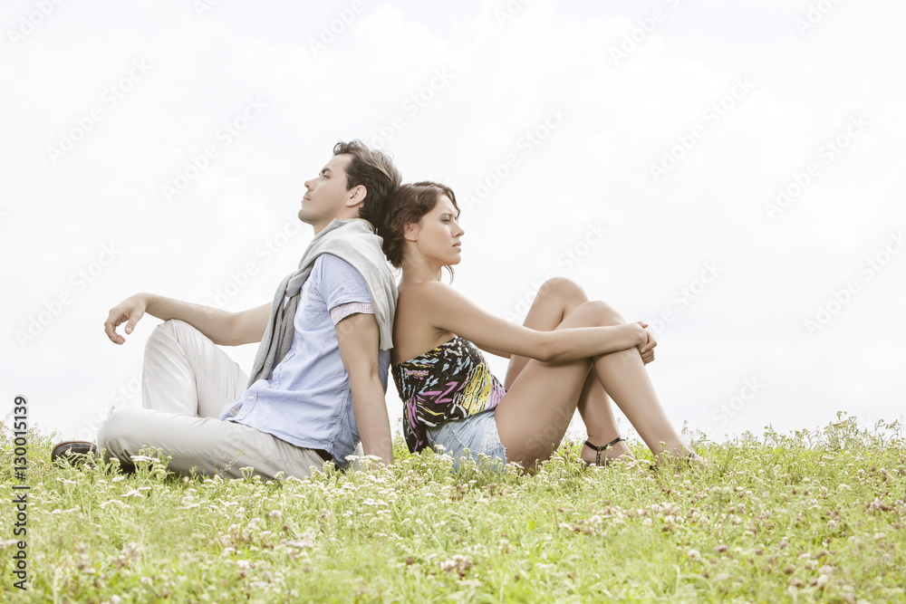 Full length of thoughtful young couple sitting back to back in park Full length of thoughtful young couple sitting back to back in park