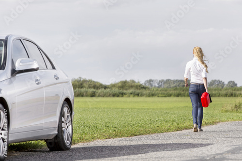 Full length rear view of woman carrying gas can leaving behind broken down car at countryside © moodboard