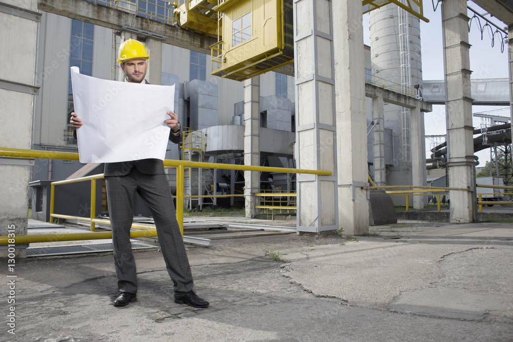 Full length of young male architect examining blueprint outside industry