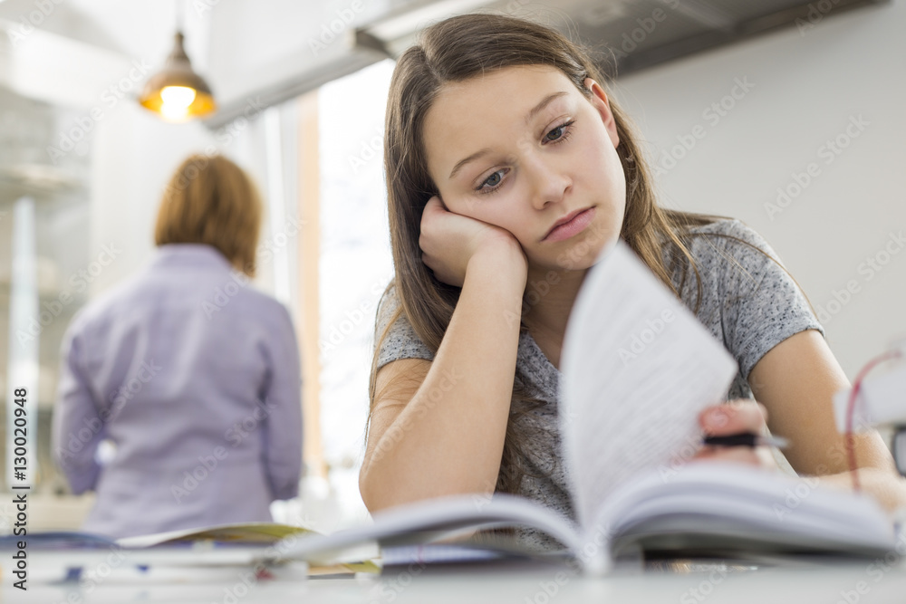Bored girl studying at table with mother standing in background Bored girl studying at table with mother standing in background
