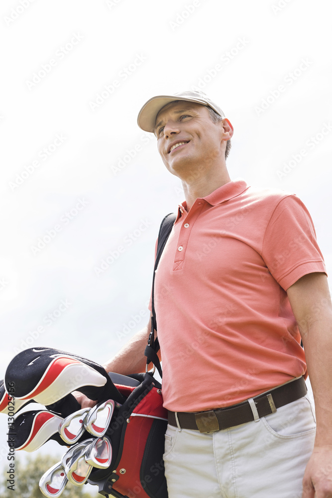 Fototapeta premium Happy middle-aged man looking away while carrying golf bag against clear sky