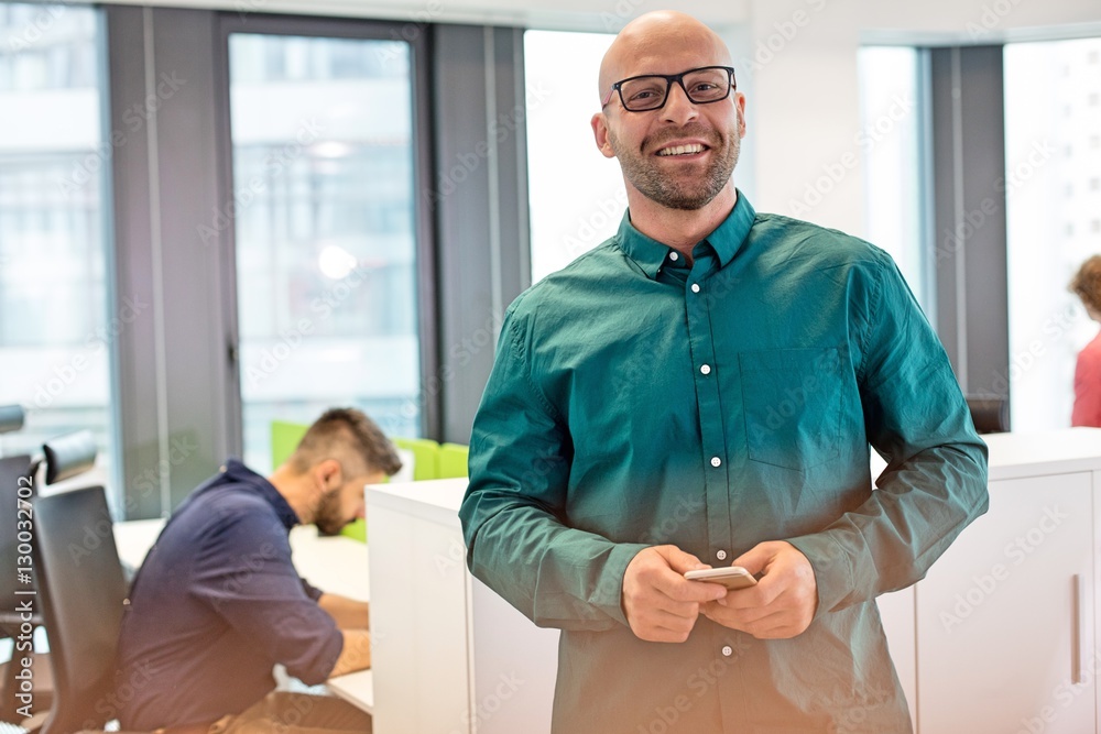 Obraz premium Portrait of confident mid adult businessman smiling with colleague in background at office