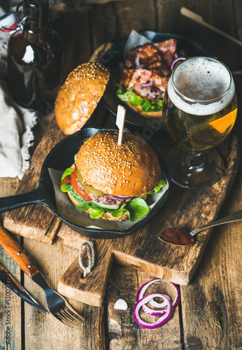 Homemade beef burgers with crispy bacon and vegetables in small black pan and glass of wheat beer on rustic serving board over shabby wooden background, top view, selective focus, vertical composition