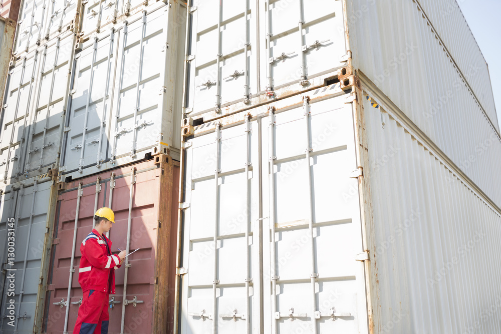 Male worker inspecting cargo containers while writing on clipboard in shipping yard