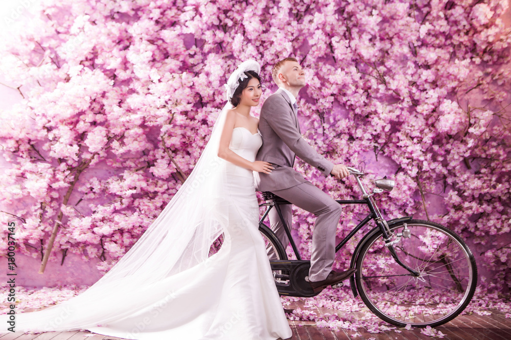 Fototapeta premium Thoughtful bride standing with bridegroom riding bicycle against pink flowers covering wall
