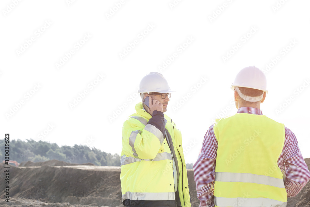 Male engineer using mobile phone while standing with colleague at construction site against clear sky