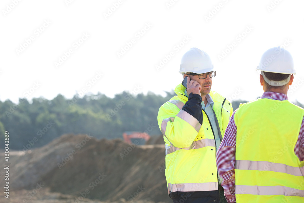 Supervisor using mobile phone while standing with colleague at construction site against clear sky Supervisor using mobile phone while standing with colleague at construction site against clear sky