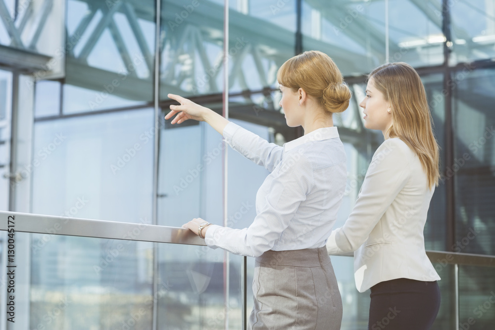 Businesswoman showing something to female colleague from glass wall in office Businesswoman showing something to female colleague from glass wall in office