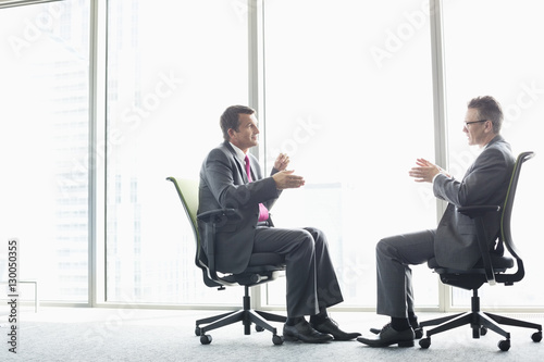 Full-length side view of businessmen discussing while sitting on office chairs by window