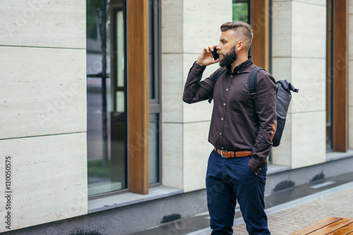 Young bearded businessman in a brown shirt with a backpack standing outdoors and talking on his cellphone. In the background is a modern building. Man using digital gadget.