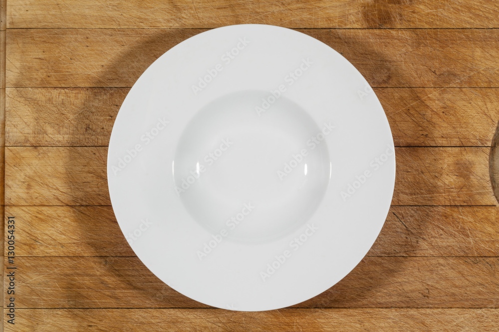 Deep white small soup plate with wide shoulders on wooden cutting board from above Deep white small soup plate with wide shoulders on wooden cutting board from above