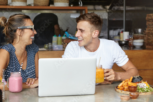 Beautiful couple having lively conversation sitting at table with laptop and food in cozy cafeteria interior, looking at each other and laughing. Young man with glass of juice talking to stylish woman
