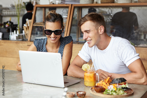 Two friends using laptop together, sitting at cafeteria. Young man wearing white t-shirt looking at screen of notebook pc with interested expression while stylish woman in shades showing him something