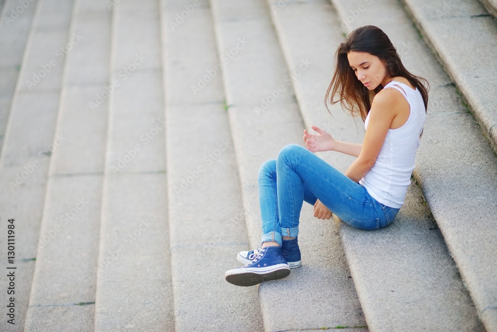 Young long-haired brunette girl in white tank top and blue jeans sitting  pensively on the steps of stone stairs. Stock Photo | Adobe Stock