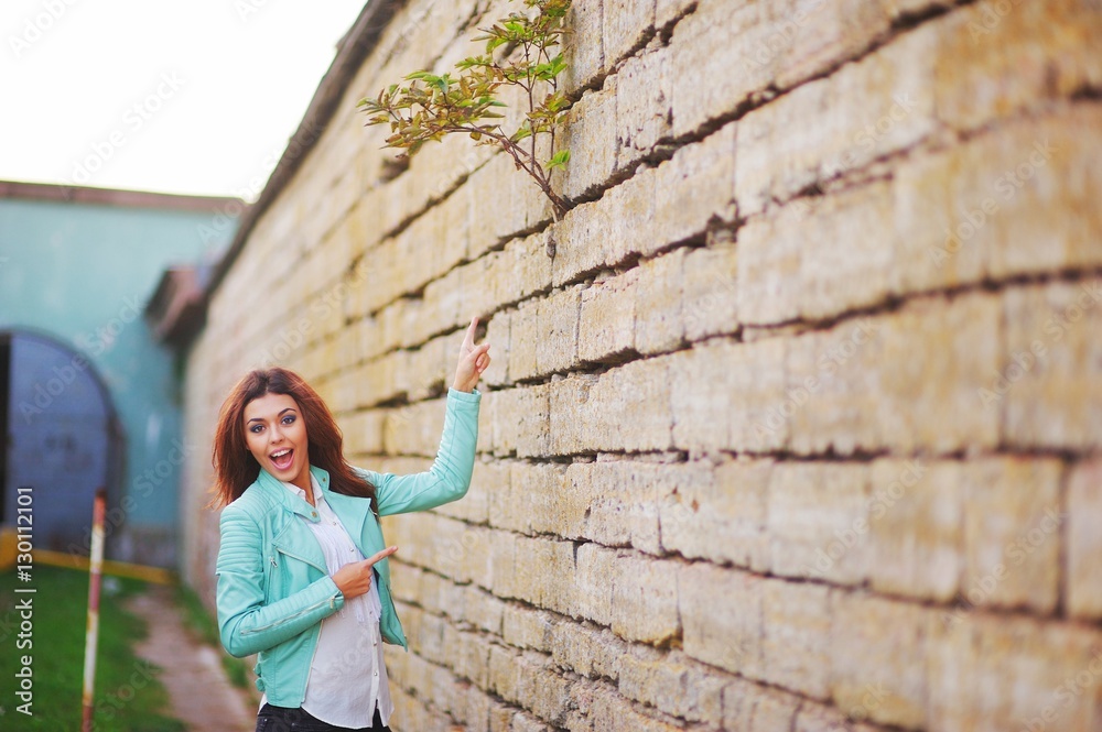 Young pretty cheerful girl standing near stone walls and delight shows on top of fence, where a young green sprout broke through a crack in masonry. The concept of life force