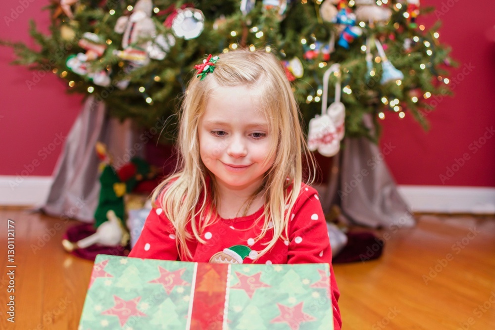 adorable child in pajamas opening christmas present in front of tree